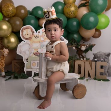 A toddler wearing a 'one' crown sits on a wooden toy bike at a jungle-themed first birthday party.