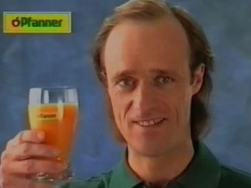 Man holding a glass of orange drink with a logo in the corner.