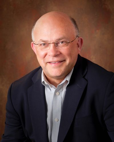 Smiling elderly man in glasses and suit jacket on brown background.