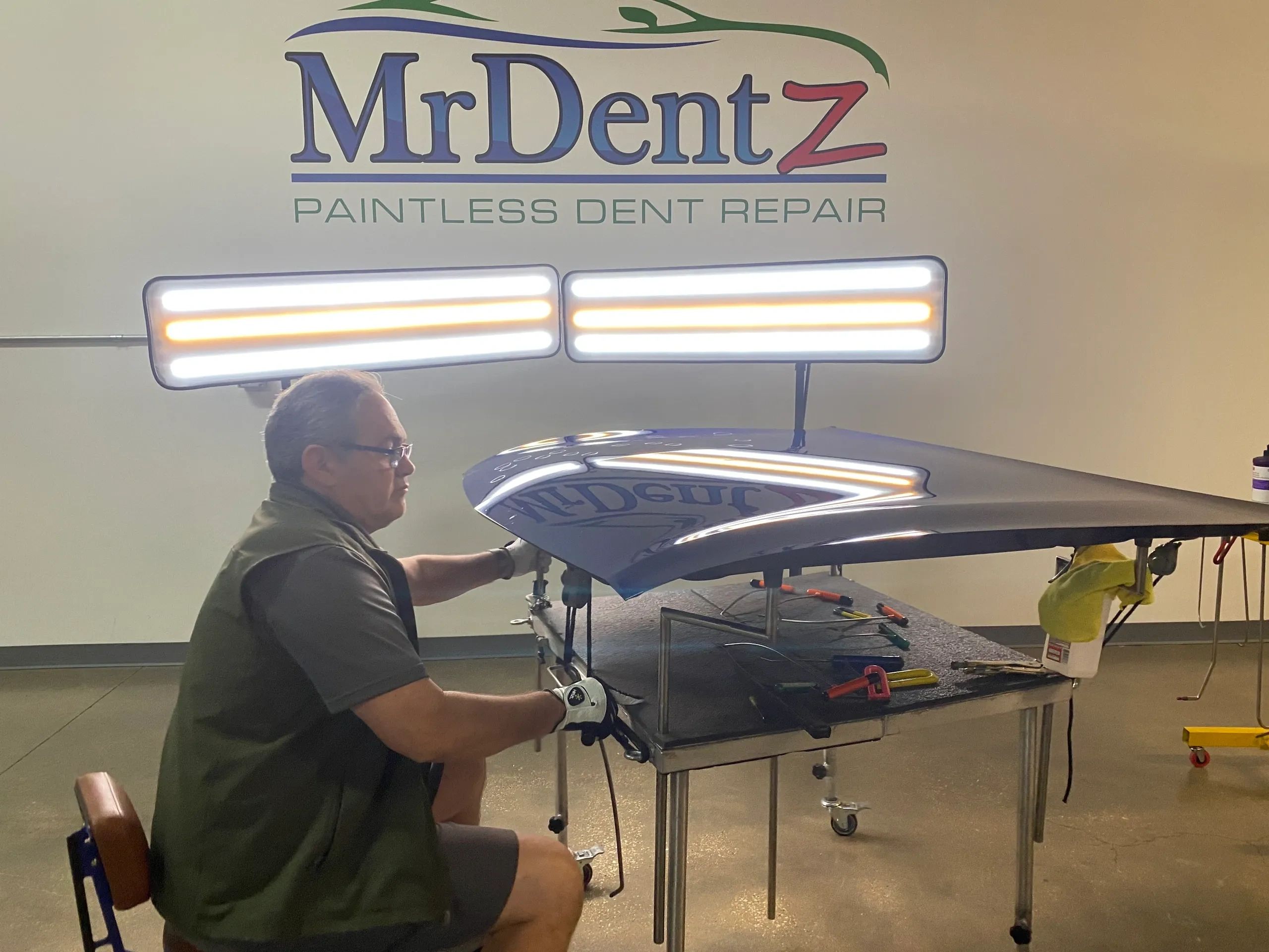 Technician performing paintless dent repair on a car hood under bright lights.