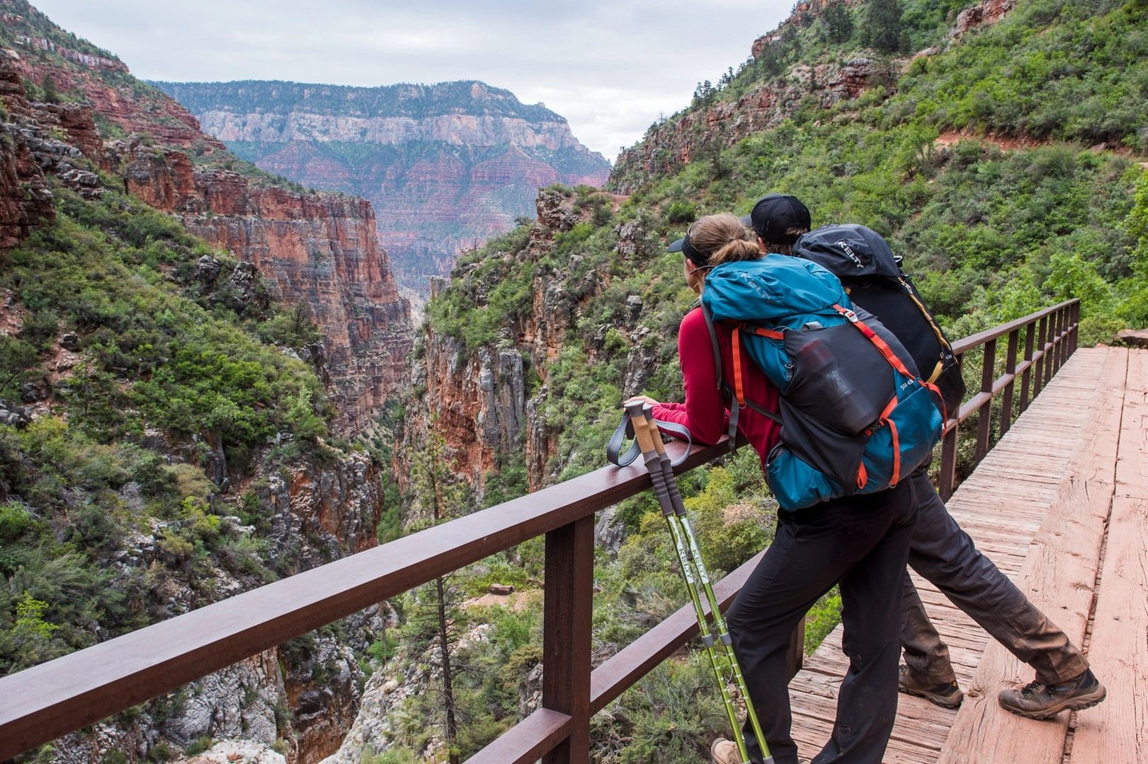 Two hikers with backpacks admire a canyon view from a wooden bridge.