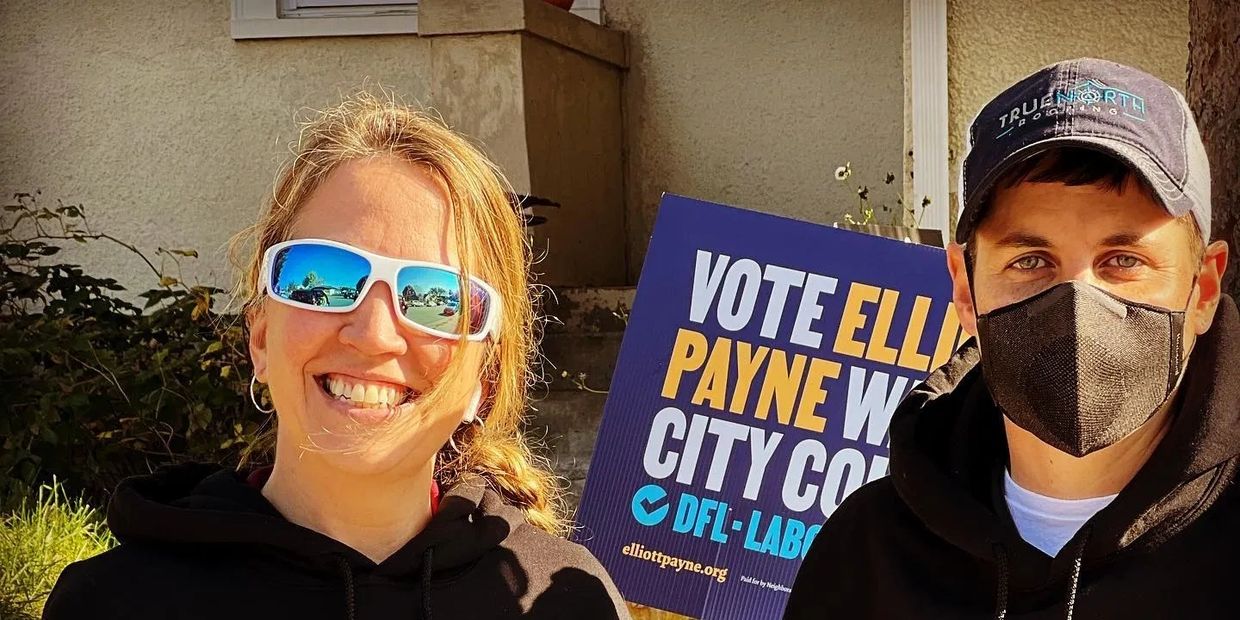 Two people stand outside with a campaign sign for Elliot Payne City Council.