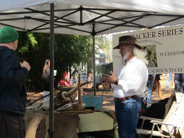 Man with long white hair and hat talks to a visitor at a historical booth.
