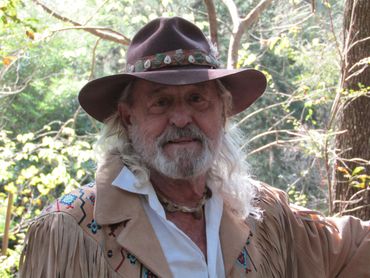 Elderly man with long white hair and beard wearing a decorated brown hat and fringed jacket outdoors.