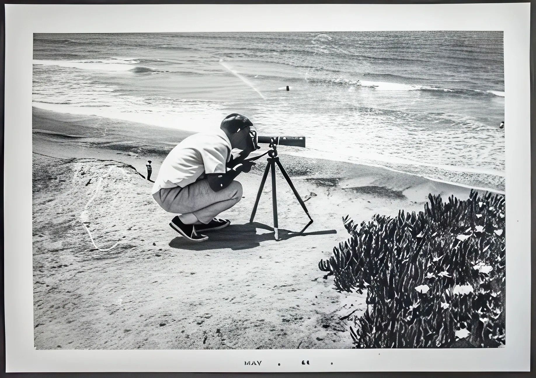Man crouches by the beach using a telescope on a tripod.