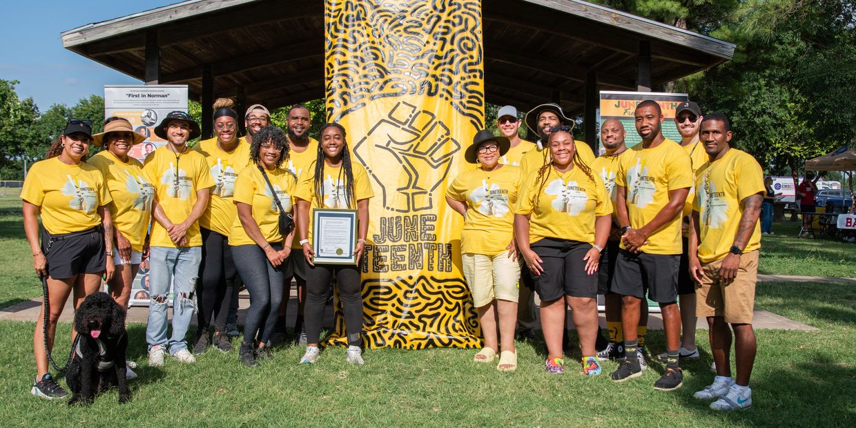 A group of 16 people stand smiling at the camera on either side of a large, yellow vertical banner. 