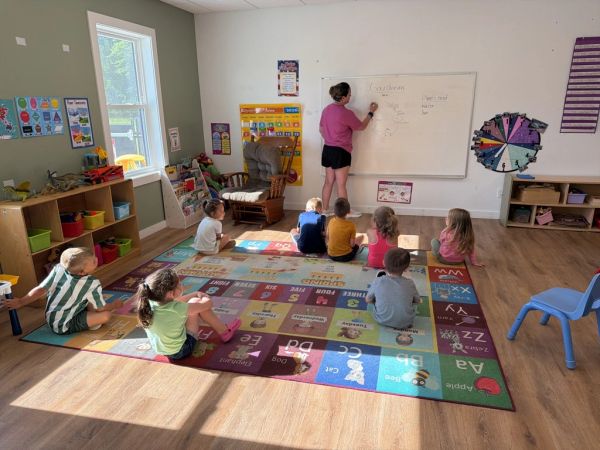 Children sitting on an alphabet rug while a teacher writes on a whiteboard in a classroom.
