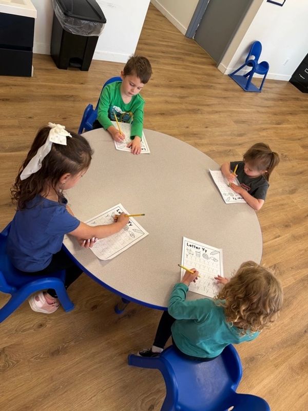 Four young children sit around a table doing worksheets with pencils.