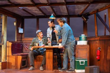 Three men in denim outfits converse intensely around a wooden table on stage.