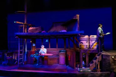 Two actors perform on a rustic wooden stage set with barrels and stairs.