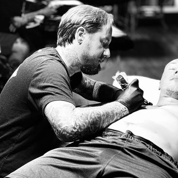 Tattoo artist working on a man's chest in a studio.