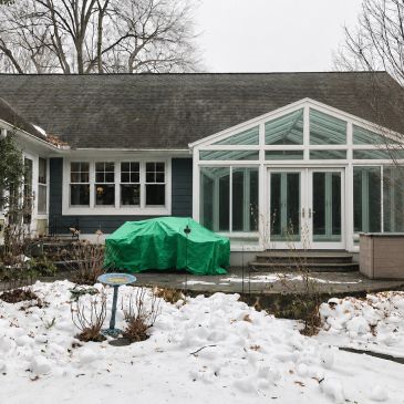 Snow-covered backyard with green-covered furniture and a glass sunroom.