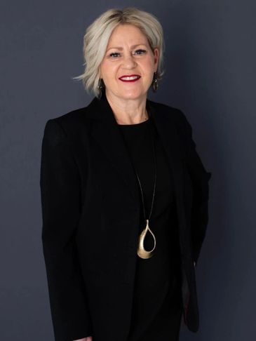 Confident woman in black attire with a gold pendant necklace against a dark background.