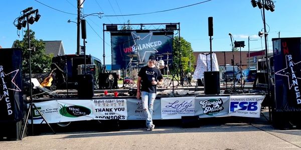 Man standing in front of an outdoor stage with music equipment and sponsor banners.
