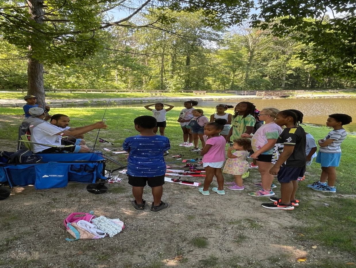 Group of children and two adults by a lake with fishing rods.