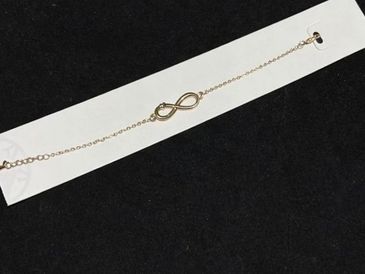 Delicate gold infinity bracelet on a white card against a black background.