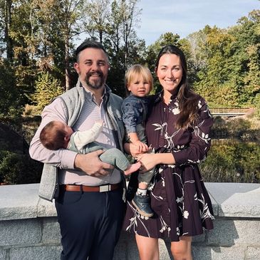 A happy family of four posing outdoors by a stone railing with trees in the background.