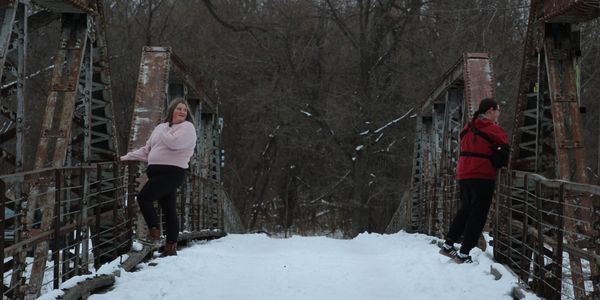 Two people stand on opposite sides of a snowy old iron bridge in winter.