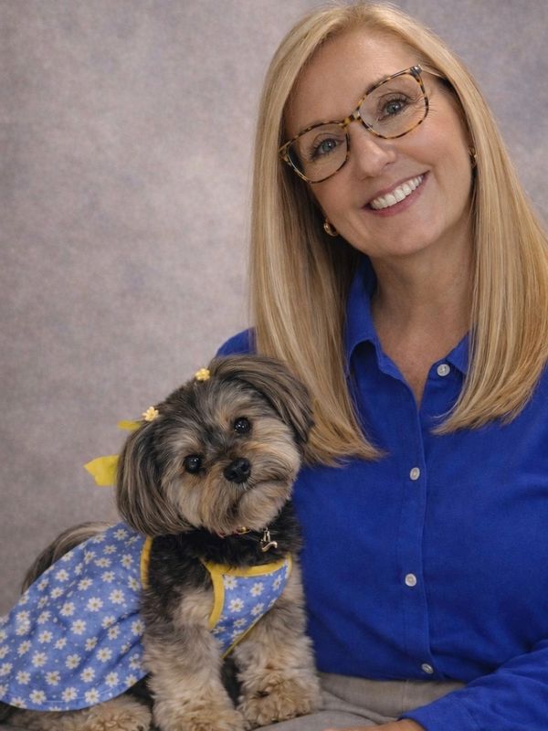 Smiling woman with glasses holding a small dog in a blue floral dress.
