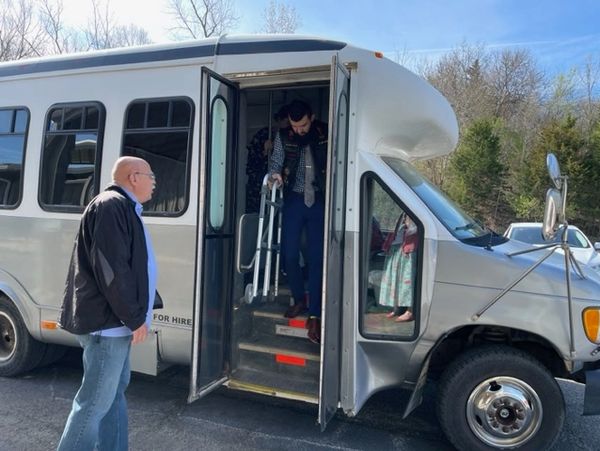 A person with a walker exits a small bus while another person waits outside.