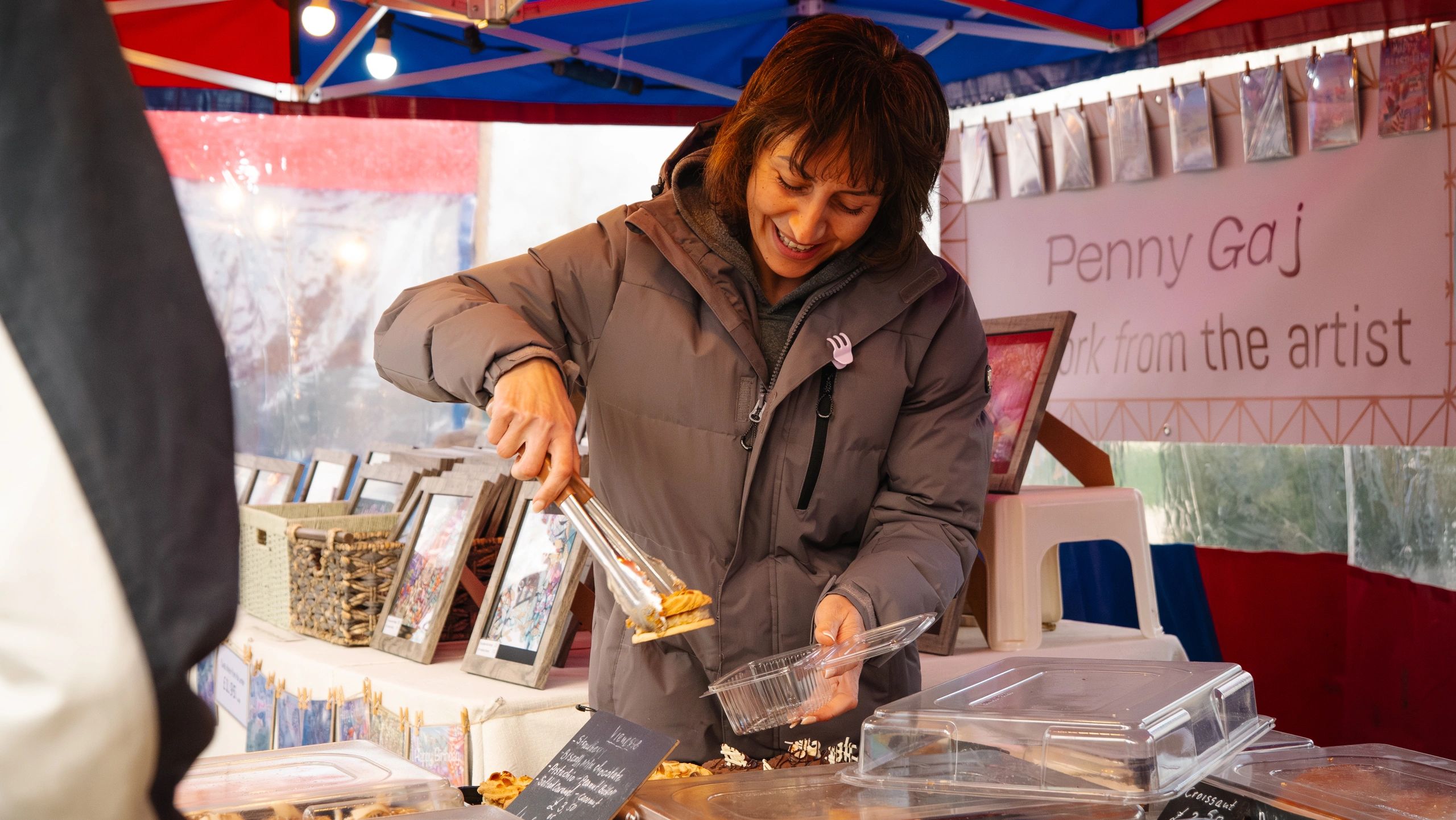 Smiling vendor serving pastries at an outdoor market stall.