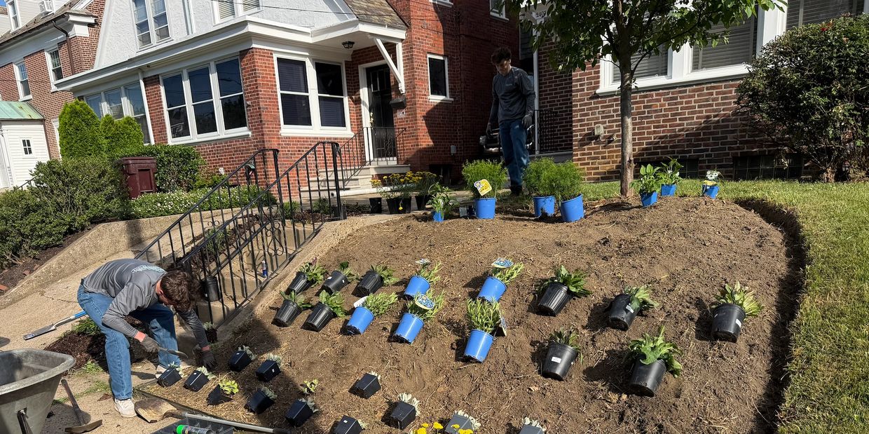 Two people planting in a garden bed in front of a brick house.