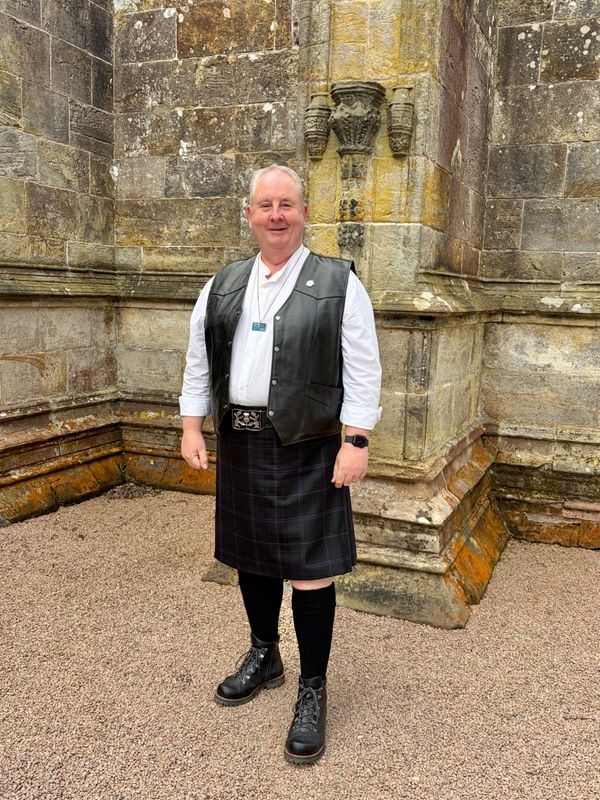 Man wearing a black kilt, leather vest, and boots standing by a stone wall.