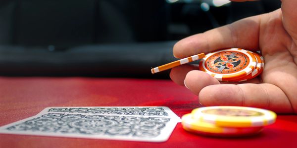 Hand holding poker chips and a cigarette on a red table with playing cards.