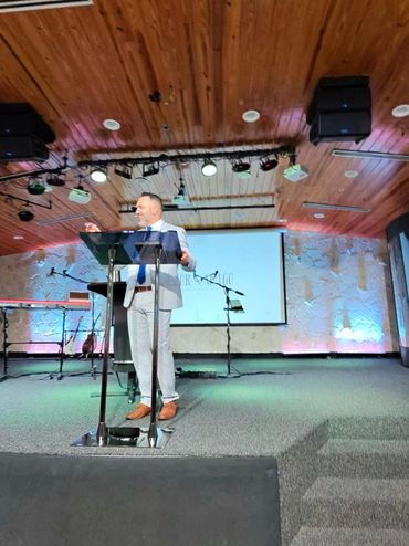 Man in a suit speaking at a podium on a stage with musical instruments in the background.