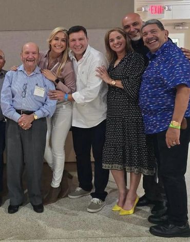 A cheerful group of six people posing indoors, smiling and embracing each other.