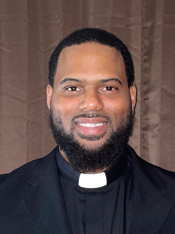 Smiling man wearing a black clerical collar shirt.