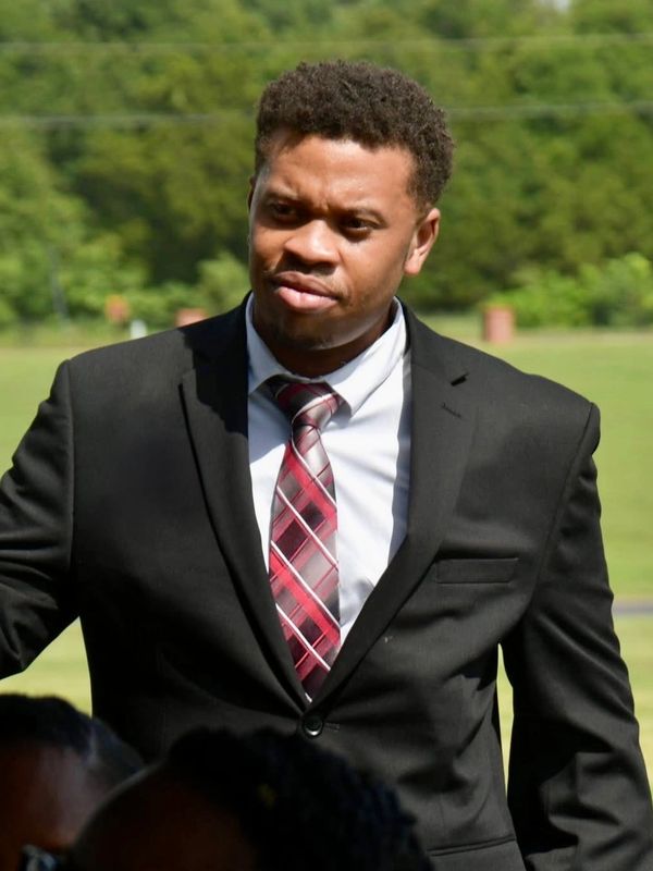 Man in a black suit with a red plaid tie outdoors.