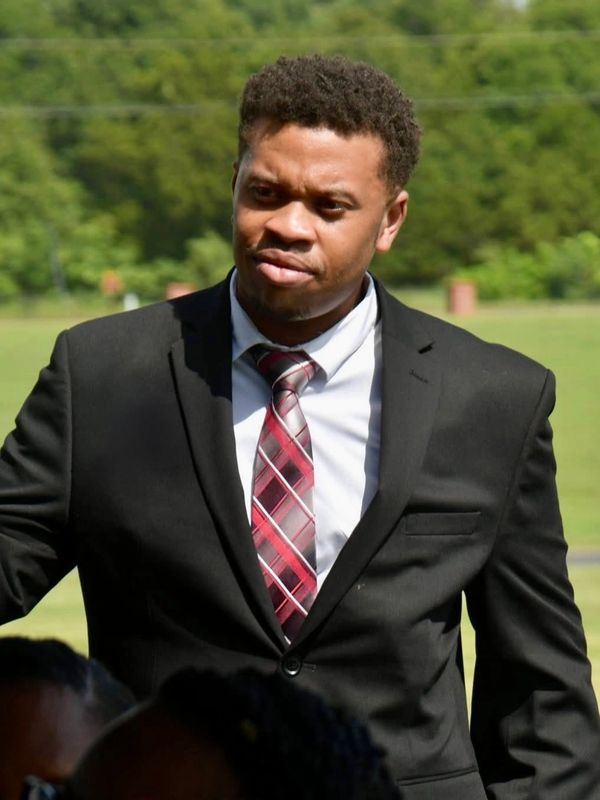 Man in a black suit with a red plaid tie outdoors.