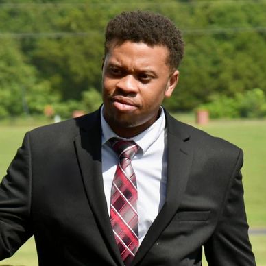 Man in black suit and red plaid tie outdoors.
