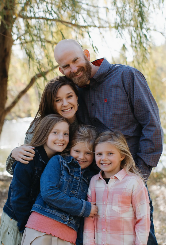 Happy family of five posing outdoors with smiles under a tree.