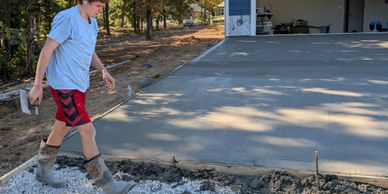 A worker walks on gravel near freshly poured concrete slab in a sunny outdoor setting.