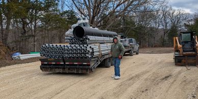 Man standing next to a truck loaded with pipes on a dirt road.