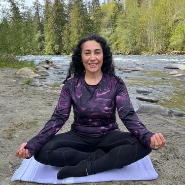 Lady sitting cross-legged beside and river in a meditation pose.
