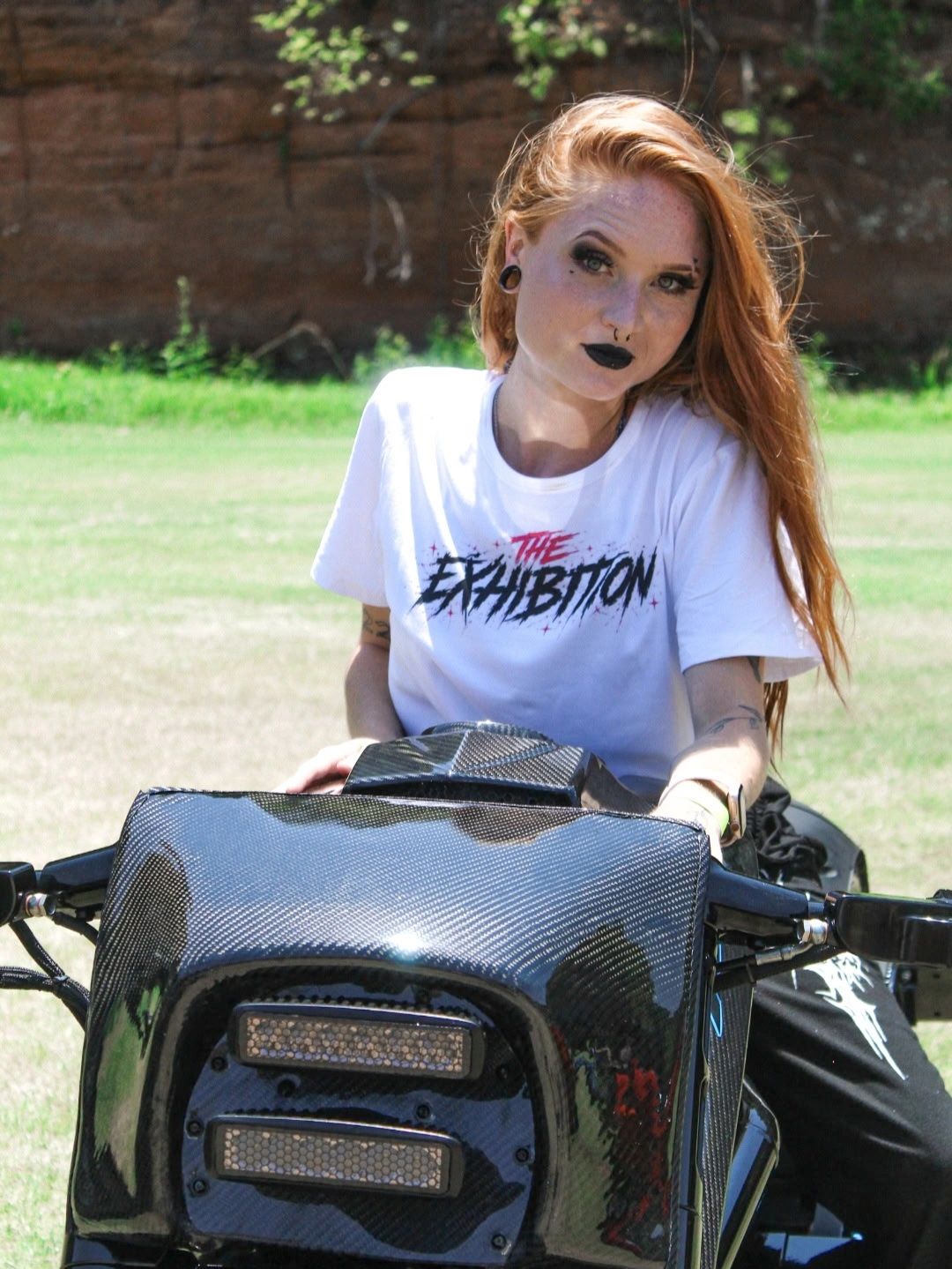 Woman with red hair on a motorcycle with "The Exhibition" shirt.