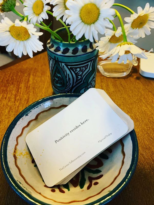 A stack of positivity cards in a decorative bowl next to a vase of daisies.