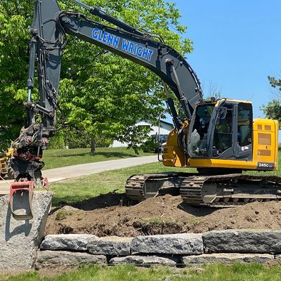 Excavator with 'Glenn Wright' on arm working on stone placement outdoors.