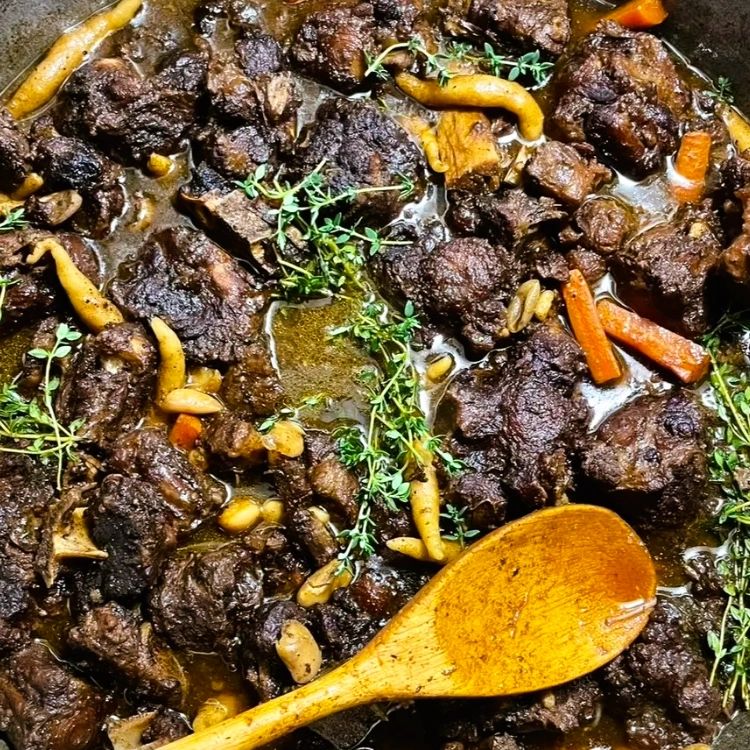Hearty Oxtail stew with carrots, herbs, spinners and a wooden spoon in a pot.