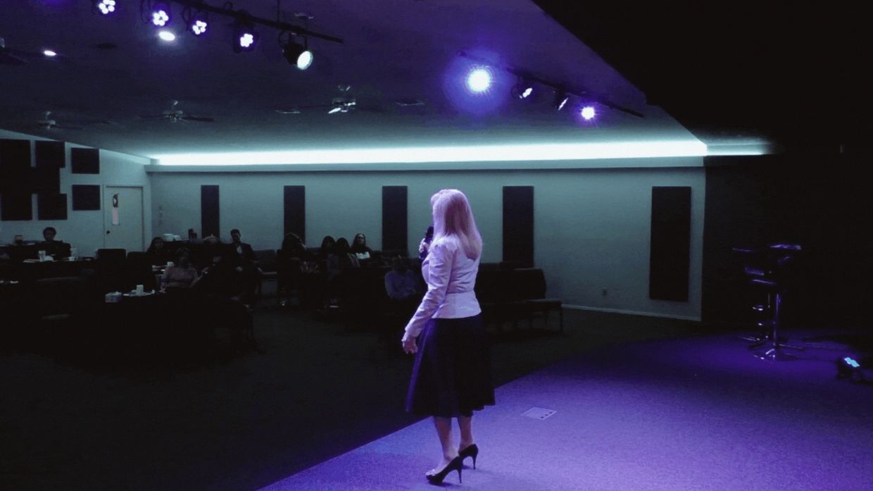 A woman in a skirt and heels speaks to an audience under purple stage lights.