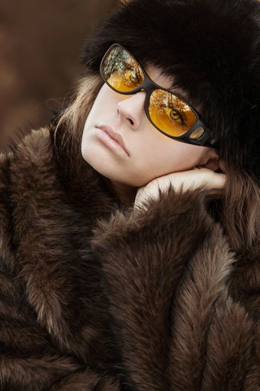 Woman in brown fur coat and hat with yellow sunglasses reflecting trees.