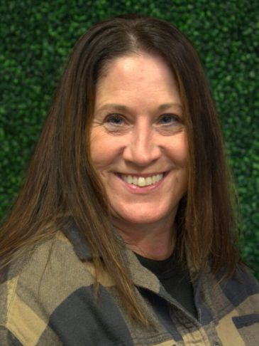 Smiling woman with long brown hair against a green leafy background.
