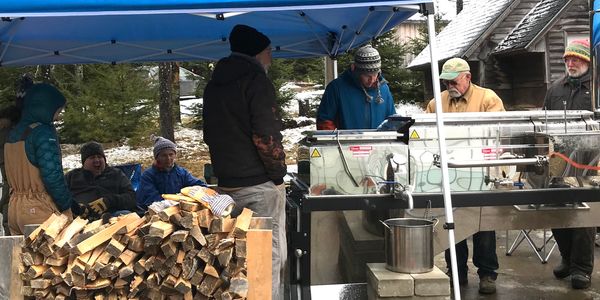 People gathered under a blue canopy with firewood and equipment outdoors.