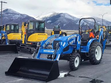 Blue New Holland tractor with loader bucket and yellow excavators in mountain backdrop.