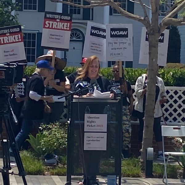 A woman speaks at a strike rally with protest signs behind her.