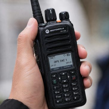 Hand holding a Motorola two-way radio with a display showing OPS TAC 1.