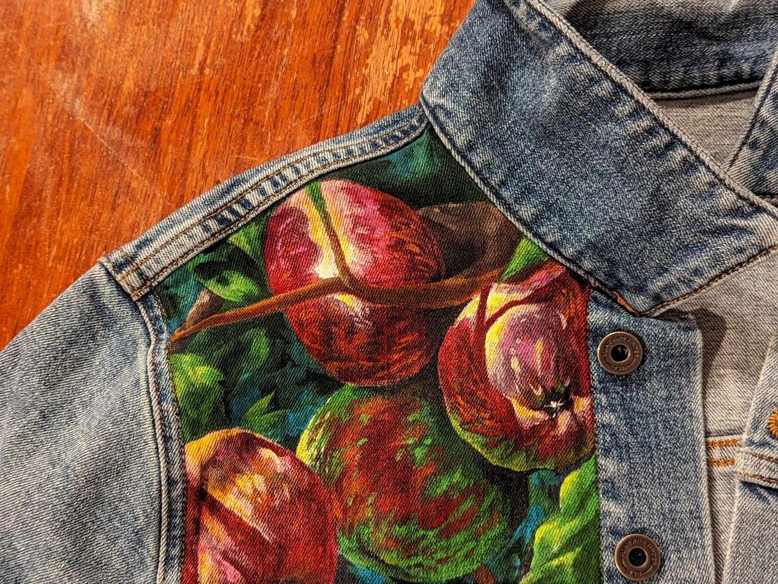 A denim jacket with one panel painted to show a close up of red apples on a tree in dappled sunlight